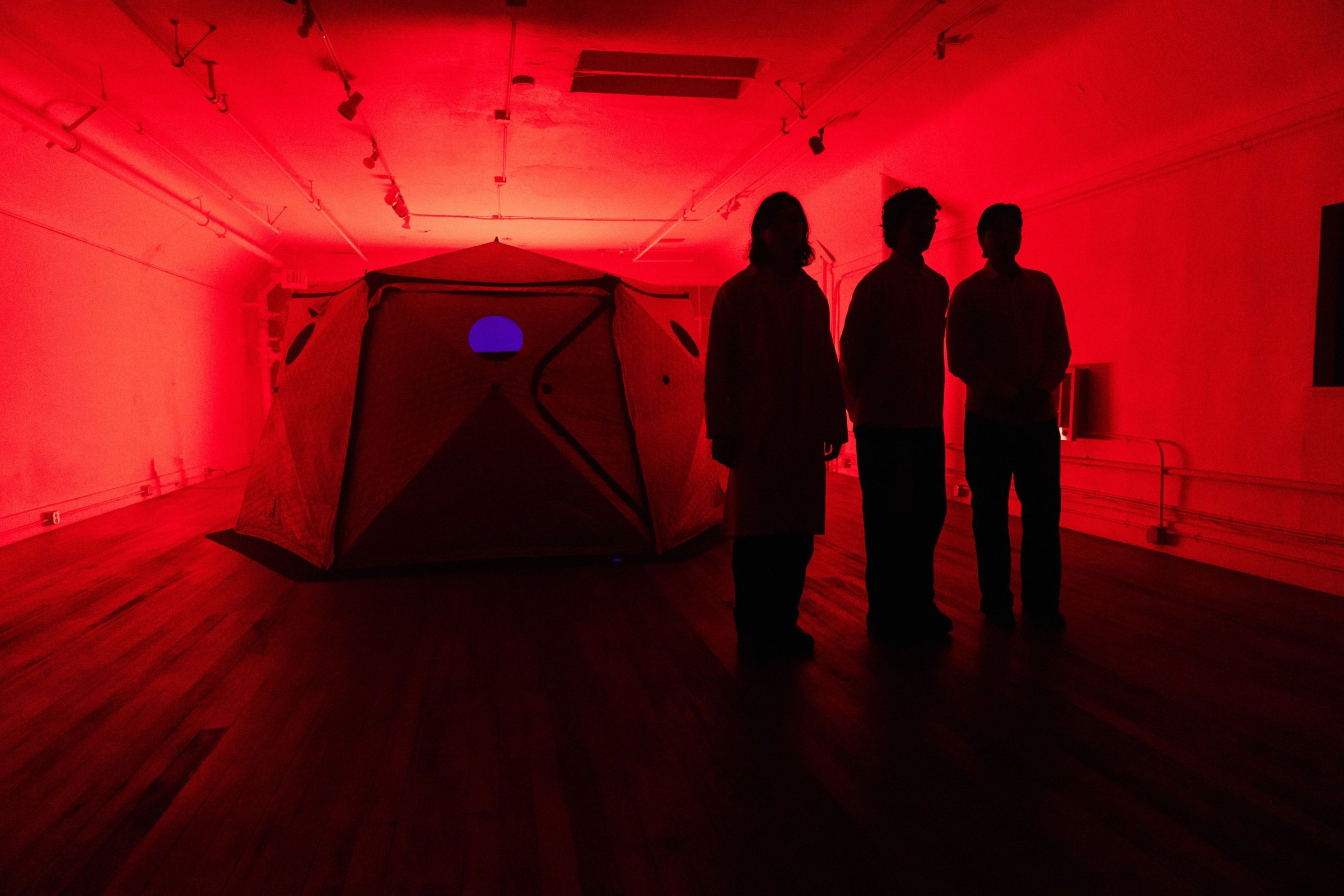 The three members of New Nostalgia standing with the HoloDome1 tent in the red-lit gallery loft