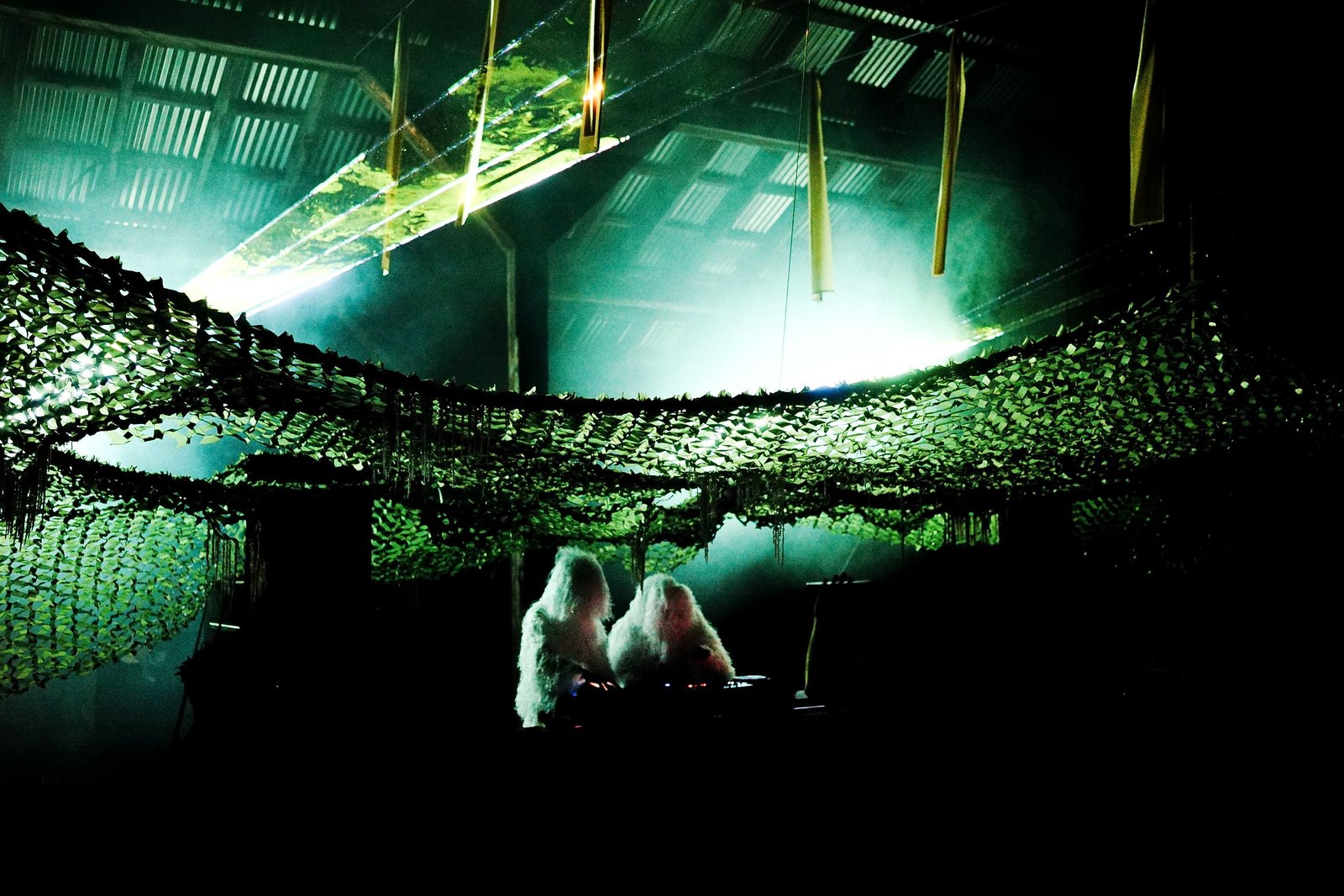 The main stage in the corrugated steel shed: strobe arrays, green net and fog with two ghillie-suited figures at the decks