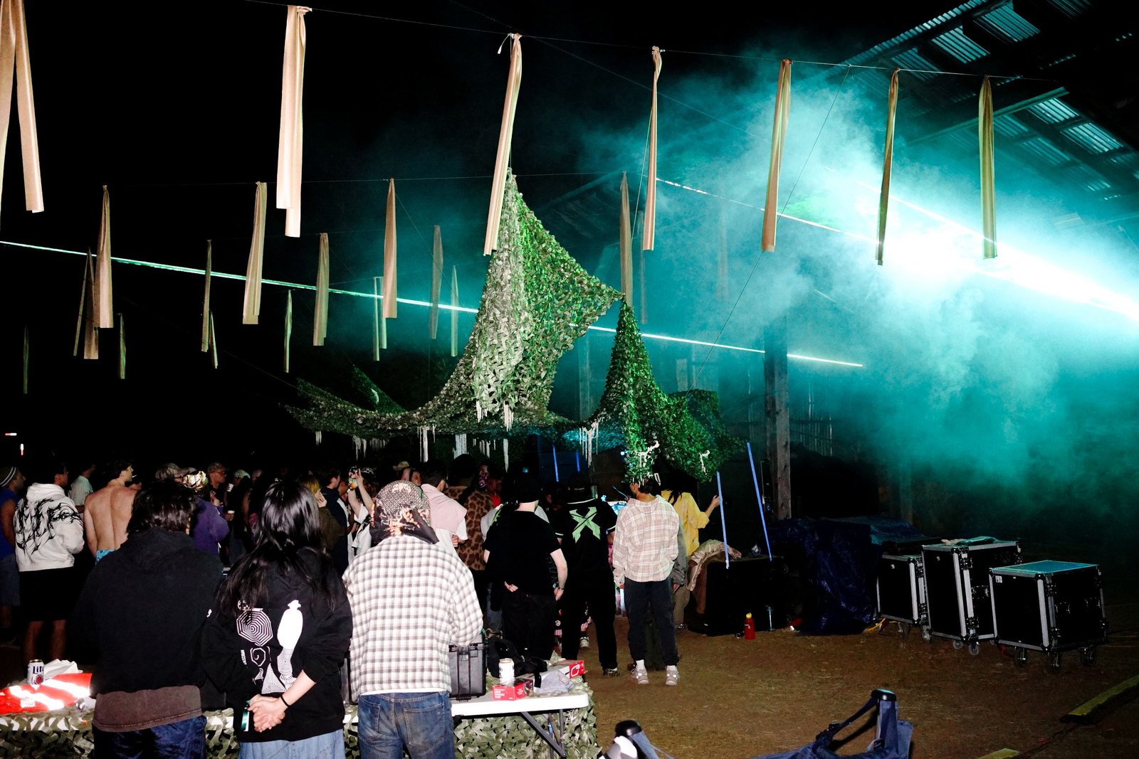 The crowd under camouflage netting at The Club, lasers cutting through fog above the corrugated shed